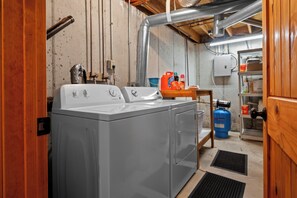 A laundry room with a front-loading washer and dryer, a blue propane tank, and various cleaning supplies on a shelf. - Nordic By Nature | Fireplace |  Playset | Fire Pit (Eden)