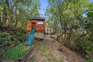A wooden playhouse with a slide, surrounded by trees and a dirt path. - Nordic By Nature | Fireplace |  Playset | Fire Pit (Eden)