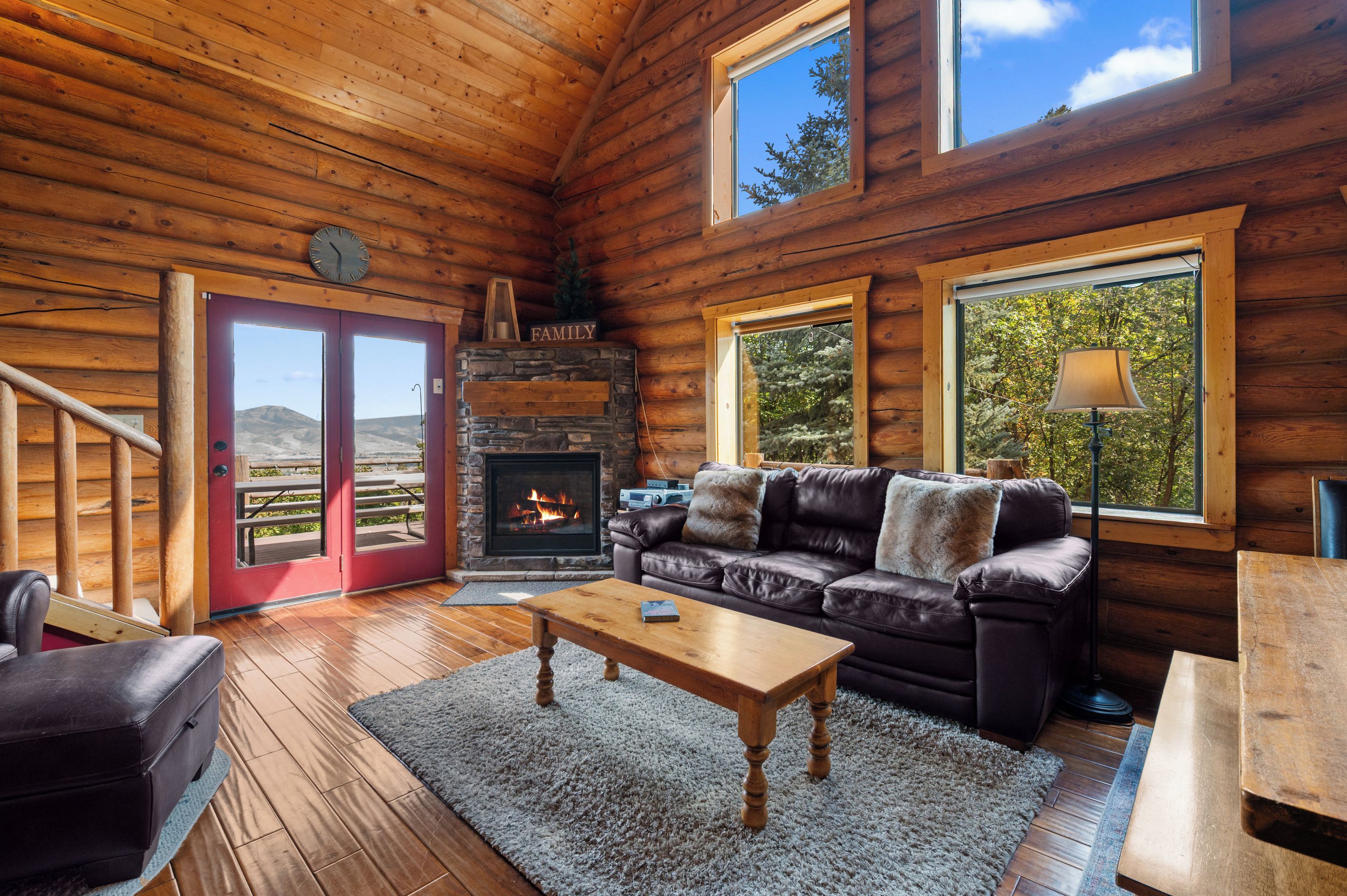 A log cabin interior with a fireplace, leather sofa, wooden flooring, and large windows.