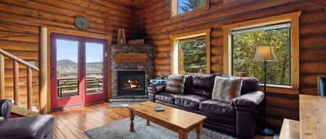 A log cabin interior with a fireplace, leather sofa, wooden flooring, and large windows.