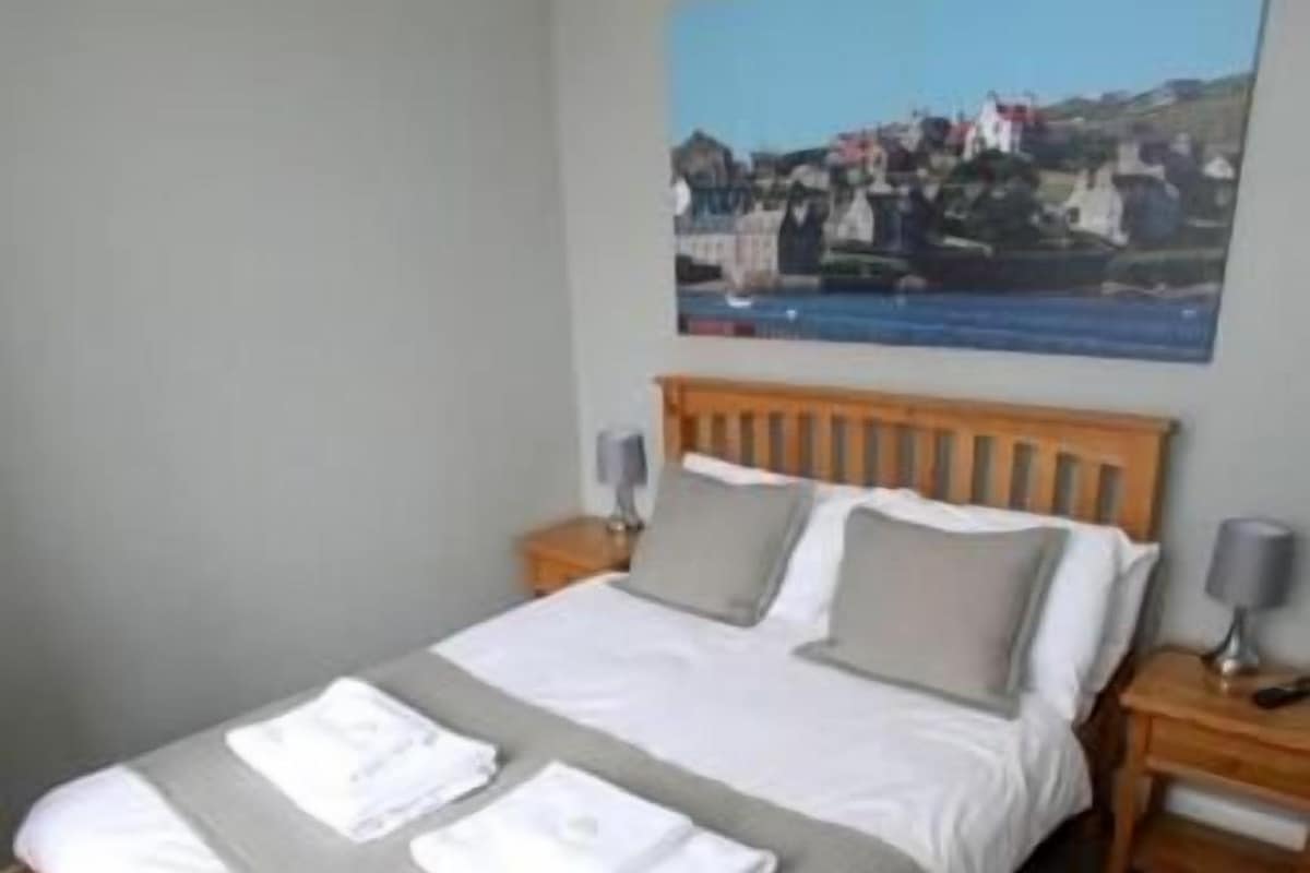 A neatly made bed with white linens and a gray blanket, wooden headboard, and a nightstand with a lamp.