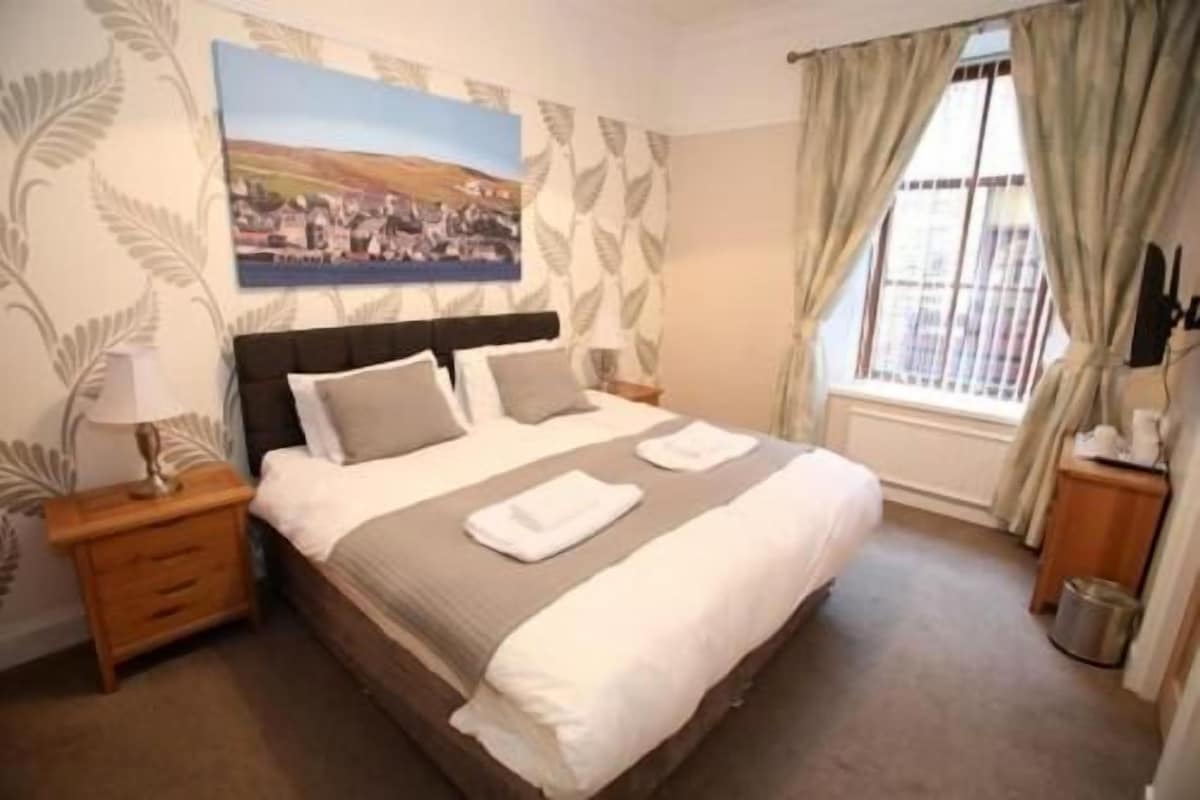 A neatly made bed with pillows and a blanket, a bedside table with a lamp, and a window with curtains.