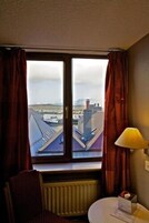 A hotel room with a large window, a chair, a table, and a lamp.