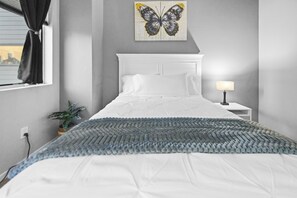 A bedroom with a bed, a nightstand, a lamp, a window with curtains, and a wall-mounted butterfly art piece. - 15-ft Ceilings · Cozy Tremont Loft · Free Parking (Cleveland)