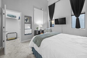 Une chambre à coucher comprenant un lit, une table de chevet, une télévision, une fenêtre avec des rideaux, une porte et une petite étagère avec quelques objets.