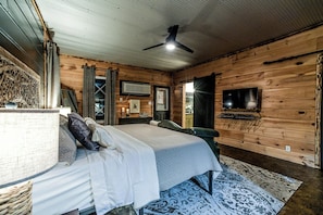 Quarto com cama, ventilador de teto, televisão presa na parede e piso de madeira.