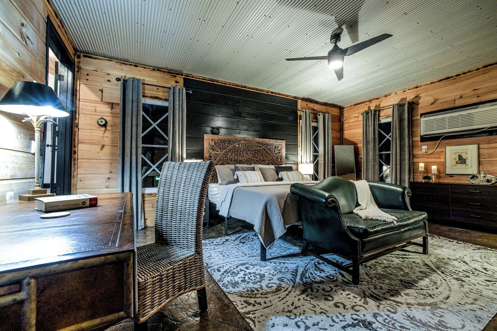 A bedroom with a bed, a desk, a chair, a ceiling fan, and a window.