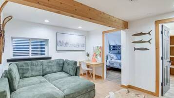 Un salon avec un canapé vert, un plafond en bois, un bureau avec une chaise, un tableau et, au fond, un lit superposé.