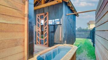 A wooden outdoor hot tub with a glass enclosure, a wooden deck, and a metal gate leading to a building with a sloped roof.