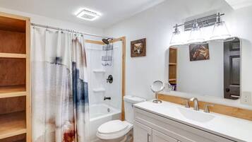 A bathroom with a shower curtain, a toilet, a sink with a mirror, and a shelf.