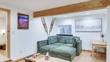 A living room with a green sofa, wooden ceiling beams, a window with blinds, and a framed picture on the wall.