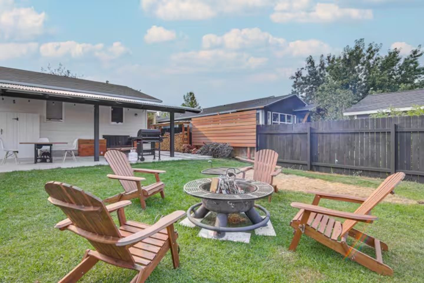 A backyard with a fire pit, four Adirondack chairs, a grill, and a covered patio area.