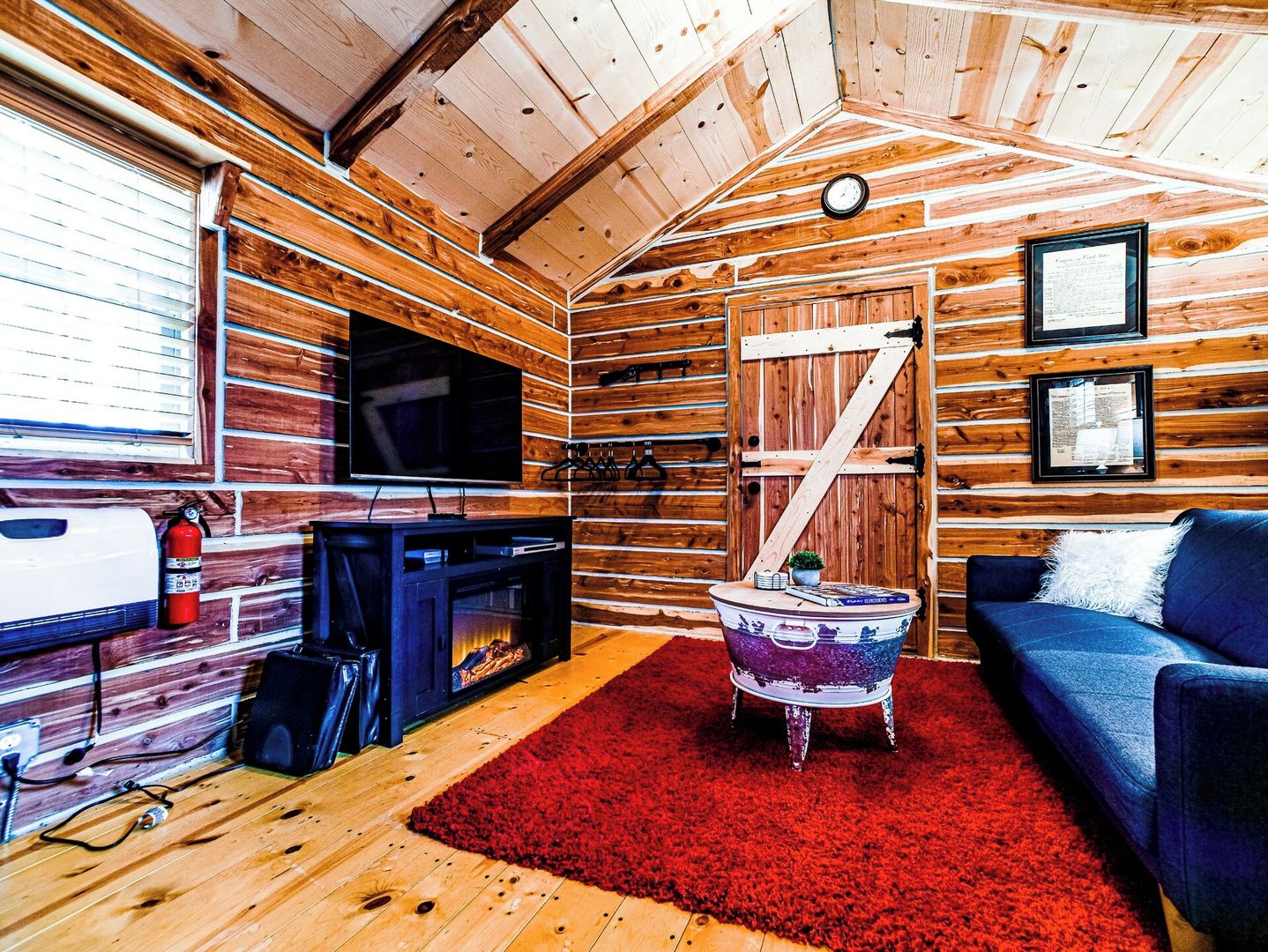 Interior de una cabaña con techo de madera, chimenea, televisor, sofá y alfombra roja.