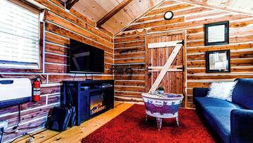 Interior de una cabaña con techo de madera, chimenea, televisor, sofá y alfombra roja.