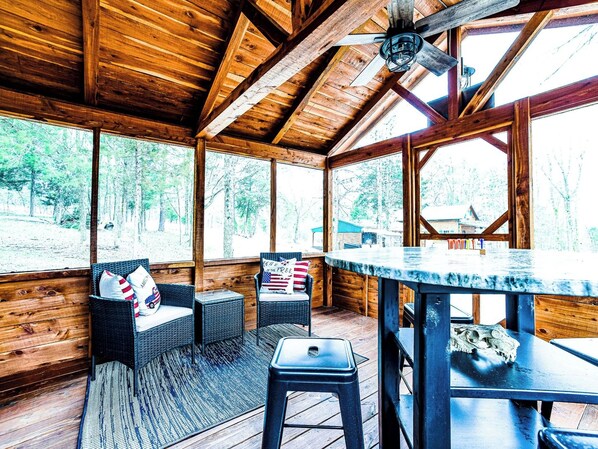 A wooden cabin interior with a high ceiling, large windows, wicker furniture, and a bar area.