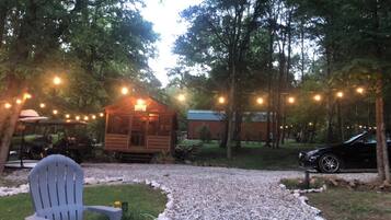 A wooden cabin with string lights, surrounded by trees and a gravel path.