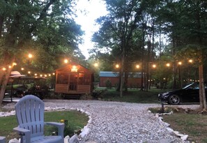 A wooden cabin with string lights, surrounded by trees and a gravel path.