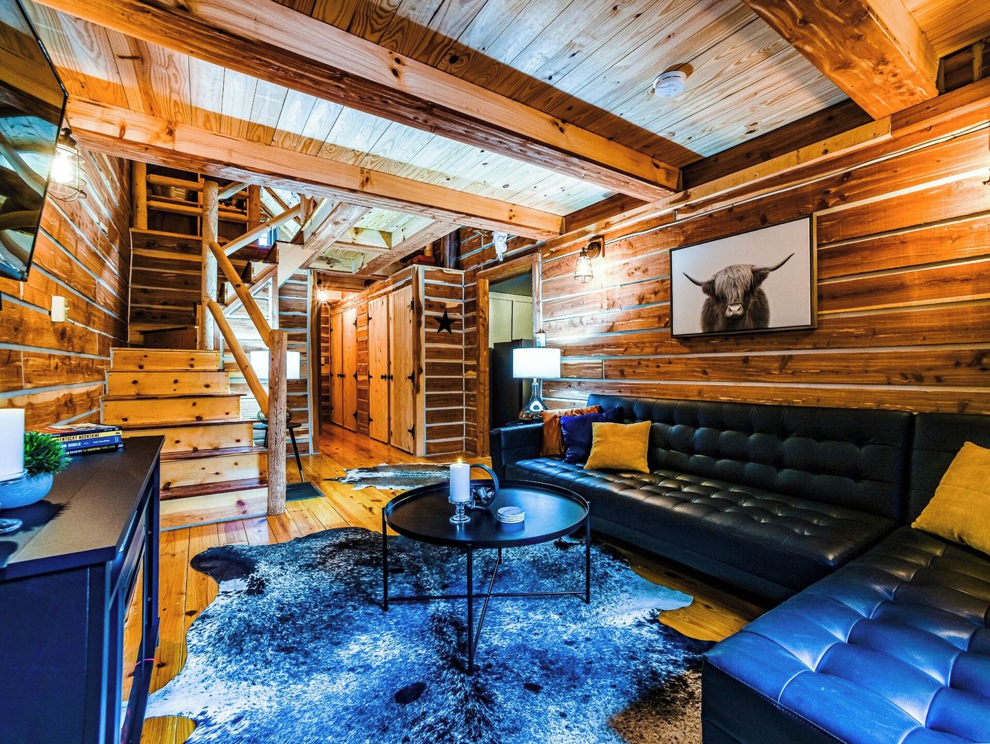 A log cabin interior with a staircase, a black leather sofa, a round coffee table, and a framed animal picture on the wall.