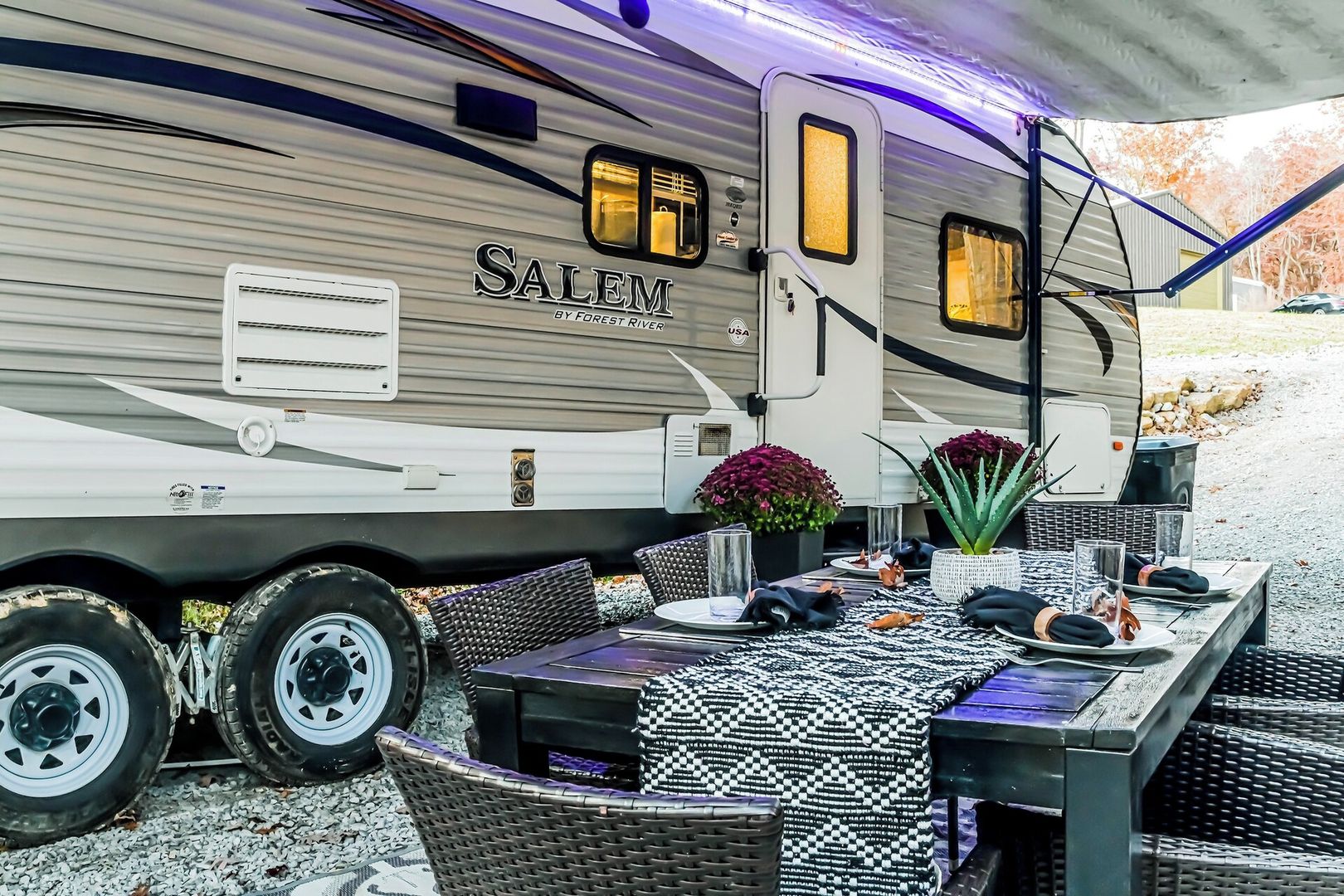 A recreational vehicle with a dining area set up outside, featuring a table with chairs, a potted plant, and a small table with a glass.