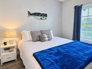 Ein ordentlich bezogenes Bett mit gemusterten Kissen, ein Nachttisch mit Lampe und ein Fenster mit Vorhängen.