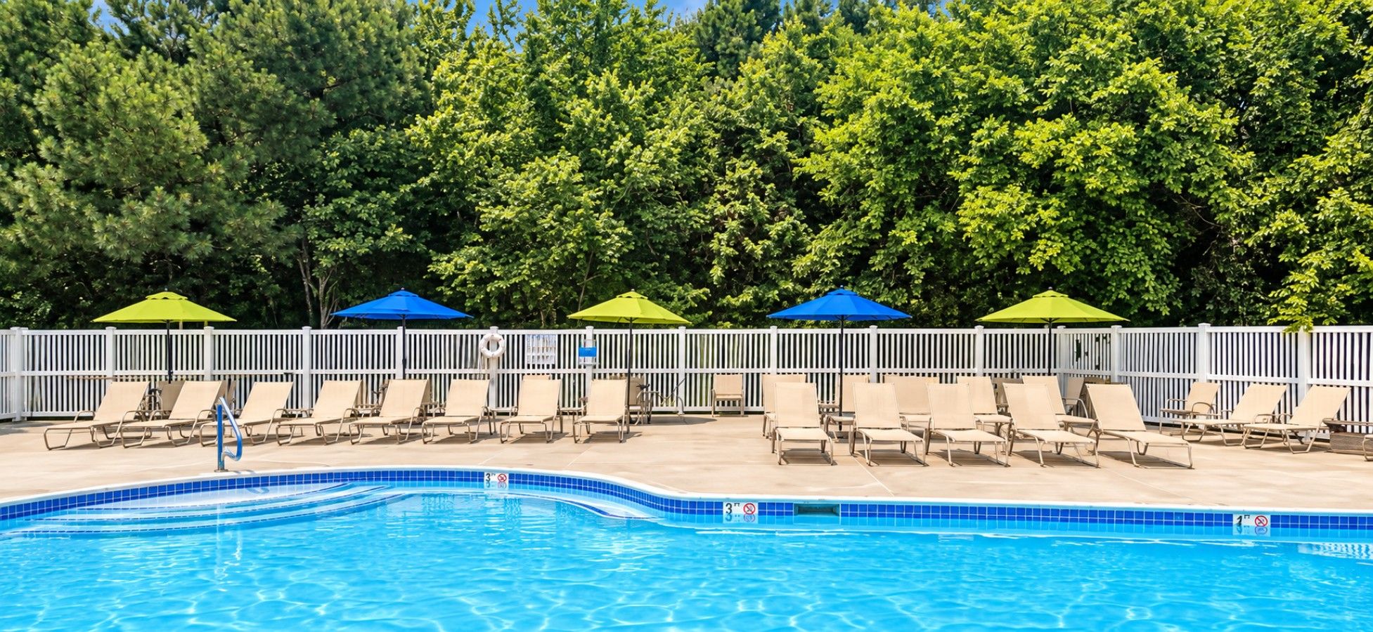 Une piscine entourée d’une clôture blanche et d’arbres, avec des chaises longues et des parasols à proximité.