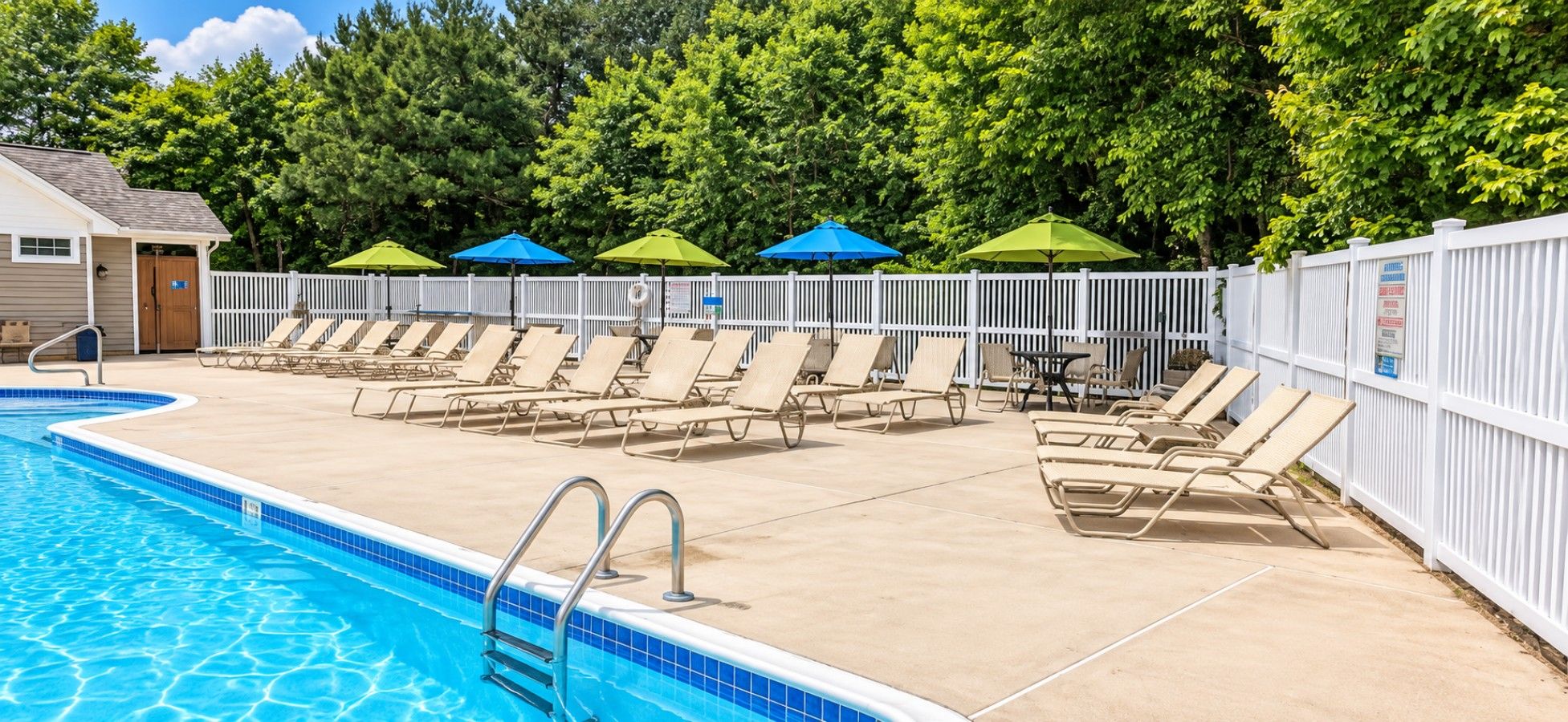 Une piscine entourée d’une clôture blanche et d’arbres, avec des chaises longues et des parasols.