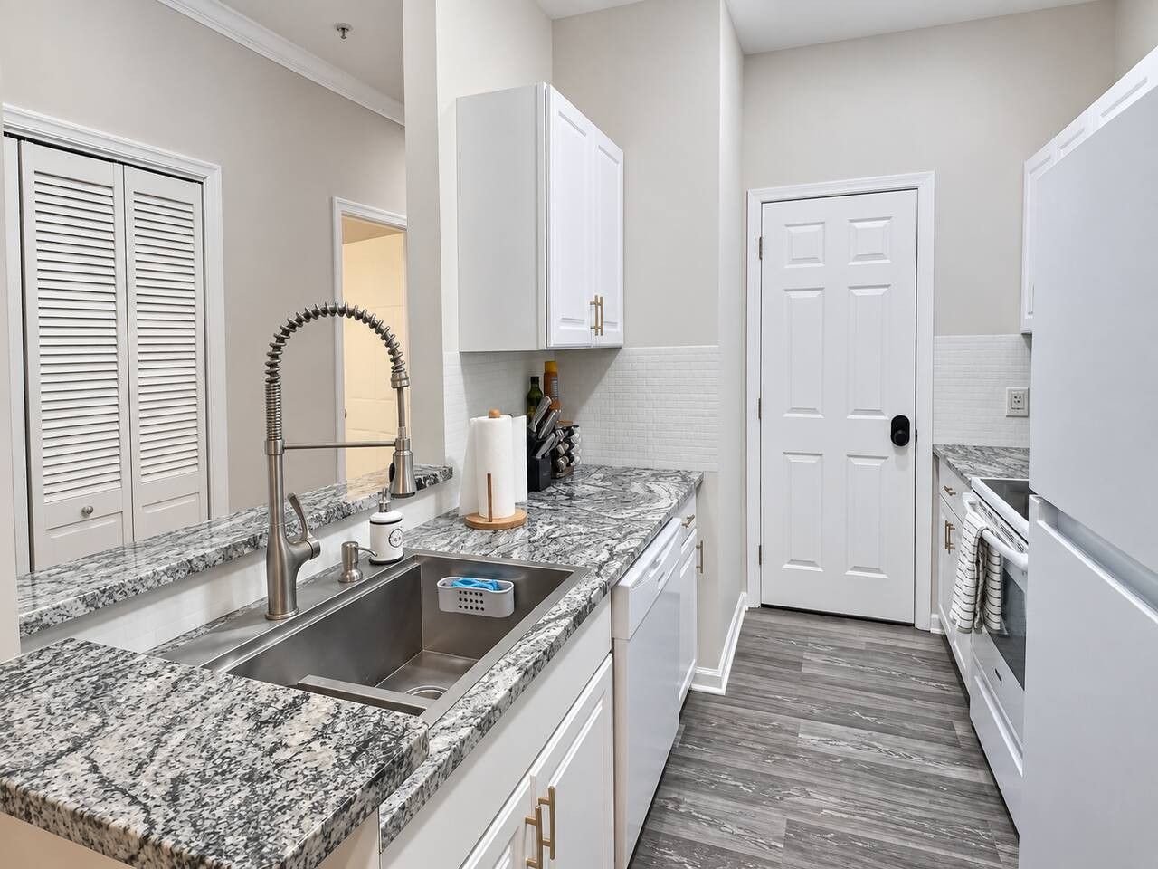 A kitchen with a granite countertop, stainless steel sink, white cabinets, and a door leading to another room.