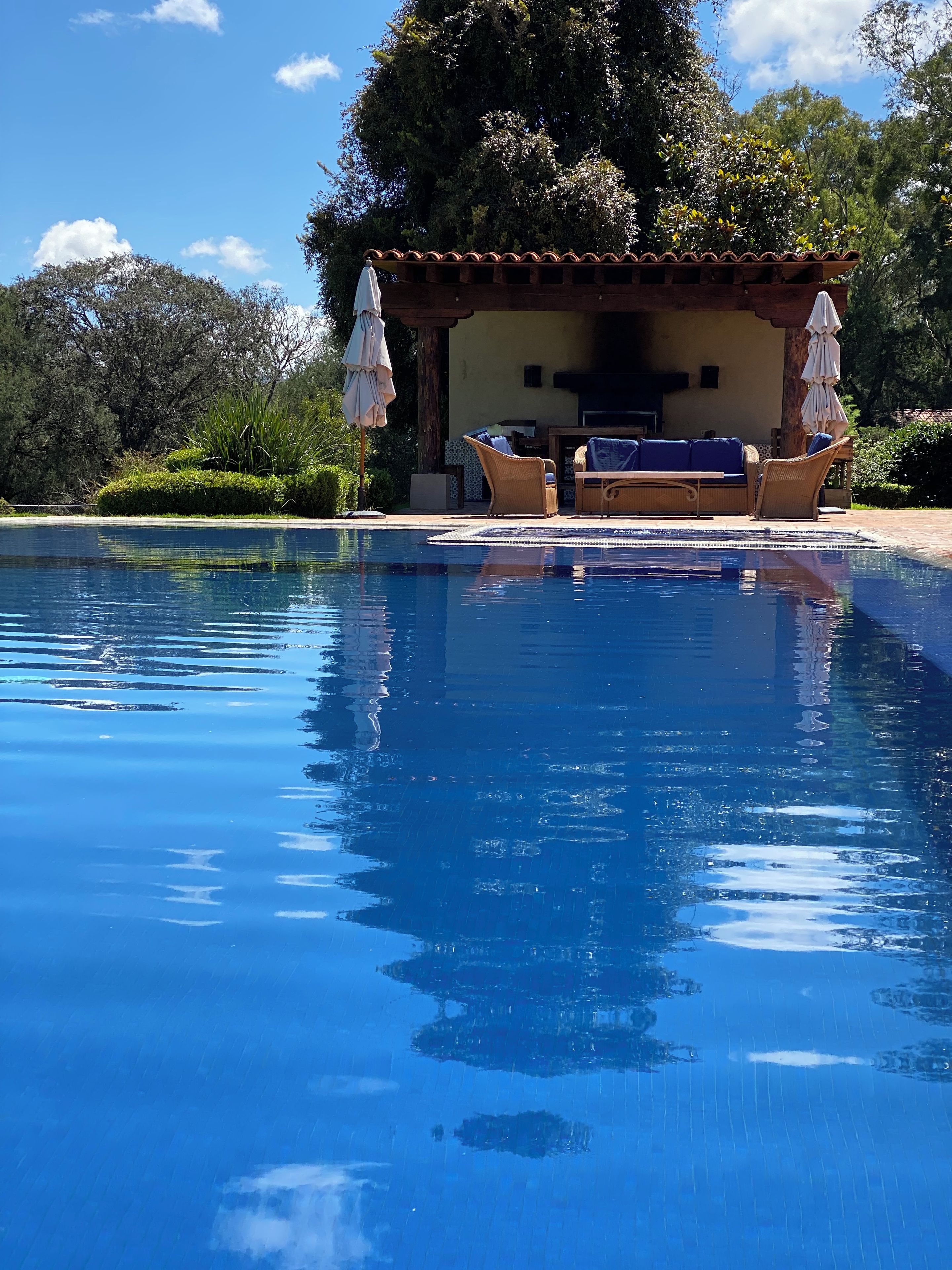 Une piscine aux eaux cristallines, bordée de chaises longues et d’ombrelles, entourée de verdure et d’un bâtiment doté d’un toit en tuiles.