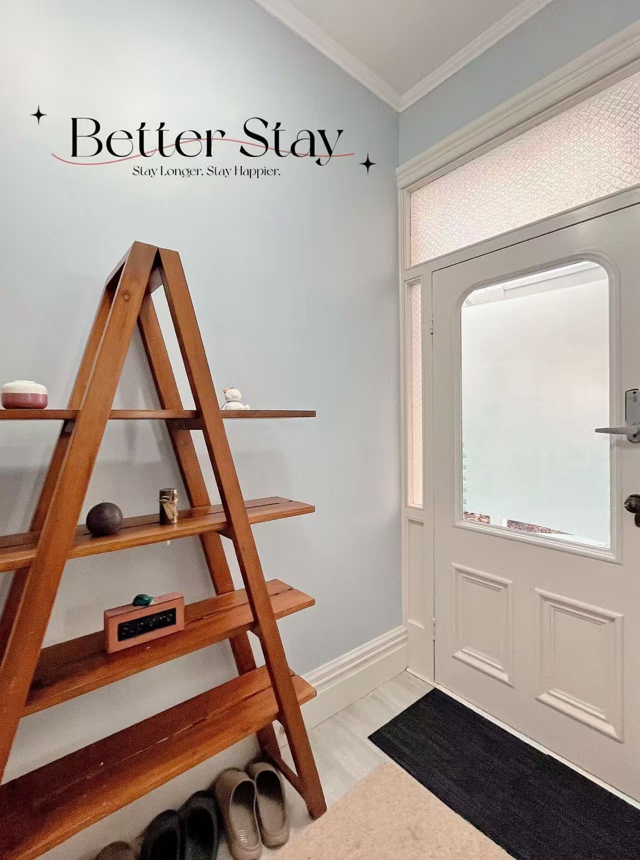 Ein Holzladderschrank mit dekorativen Gegenständen vor einer hellen Wand, eine weiße Tür mit satiniertem Glasfenster und eine schwarze Fußmatte.
