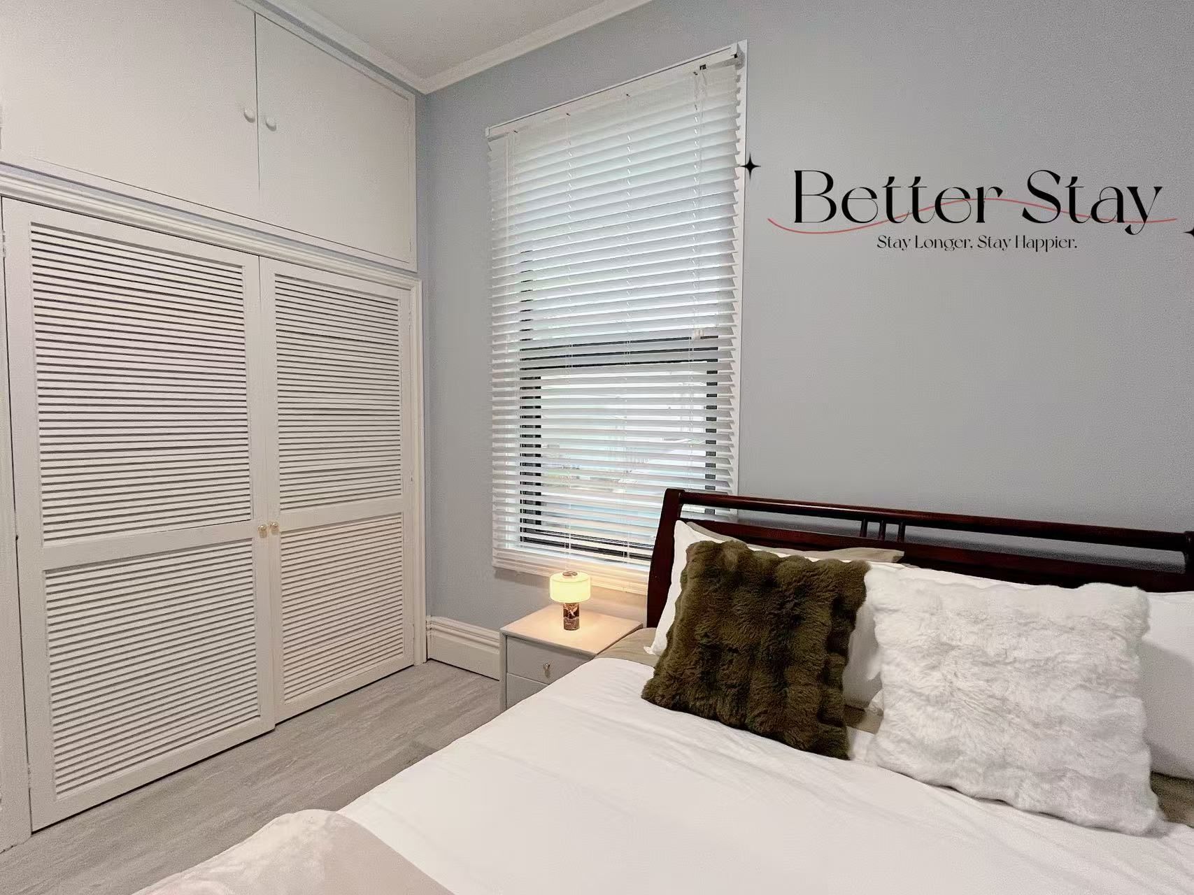 A bedroom with a bed, a nightstand, a lamp, a window with blinds, and a wardrobe.