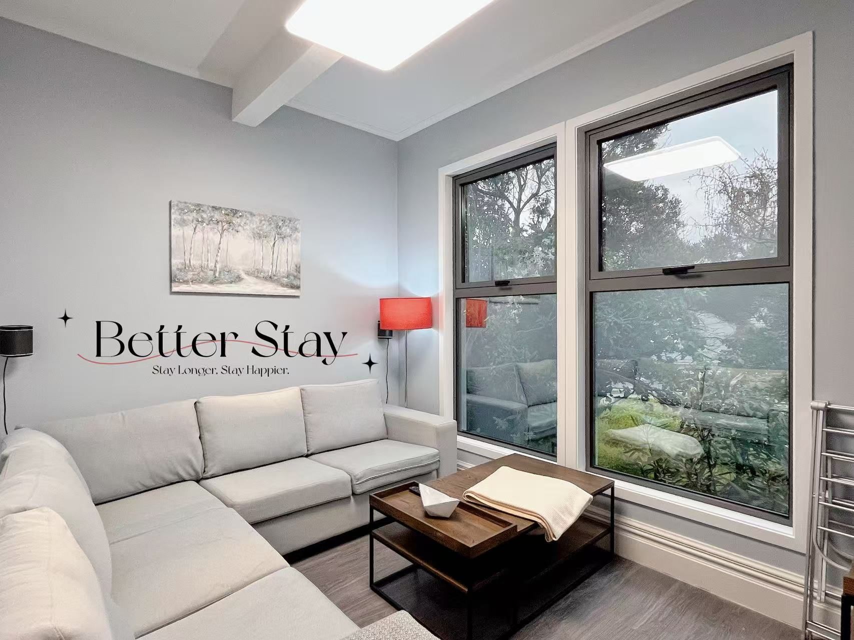 A modern living room with a grey sofa, a wooden coffee table, and a large window with a view of trees. A wall-mounted lamp and a motivational quote are also visible.