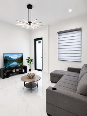 A modern living room with a flat-screen TV, a round coffee table, a grey sofa, a potted plant, and a ceiling light fixture.