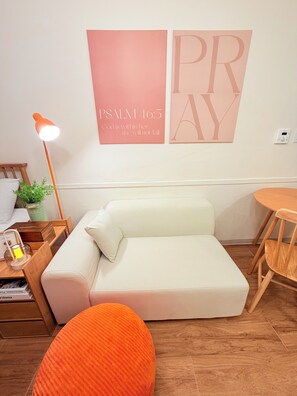 Ein gemütliches Zimmer mit einem weißen Sofa, einem kleinen orangefarbenen Hocker, einem Holznachttisch mit Lampe und einem an der Wand befestigten Bücherregal.