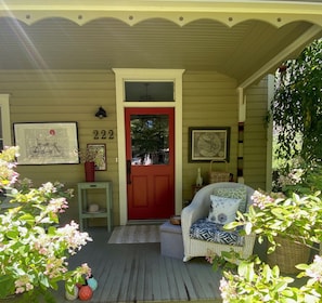 A porch with a red door, a wicker chair, and a small table with a potted plant. - Elegant 2-bedroom home in serene garden close to downtown (Paonia)