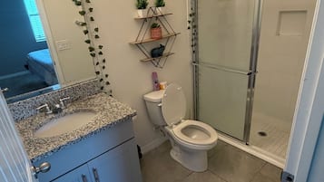 Un baño con lavamanos de encimera de granito, inodoro, ducha con puertas de vidrio y un pequeño estante con plantas.