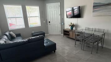 Una sala de estar con un sofá de color oscuro, una mesa de comedor con sillas, un televisor de pantalla plana montado en la pared y una puerta que da a otra habitación.