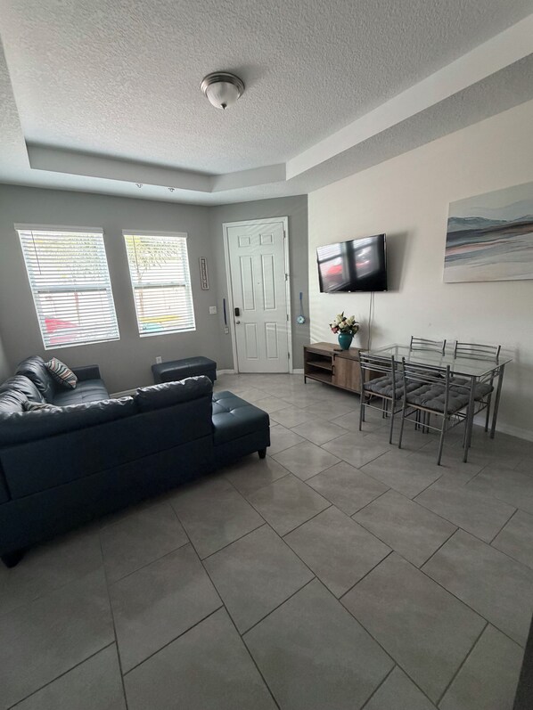 A living room with a dark-colored sofa, a dining table with chairs, a flat-screen TV mounted on the wall, and a door leading to another room. - Cozy 1-bedroom apartment in Kissimmee (Kissimmee)