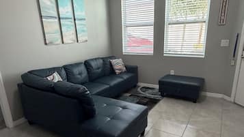 Una sala de estar con un sofá seccional, una mesa de centro y un pequeño otoman. Hay dos ventanas con persianas, una rejilla de ventilación en el techo y un cartel de bienvenida en la pared.