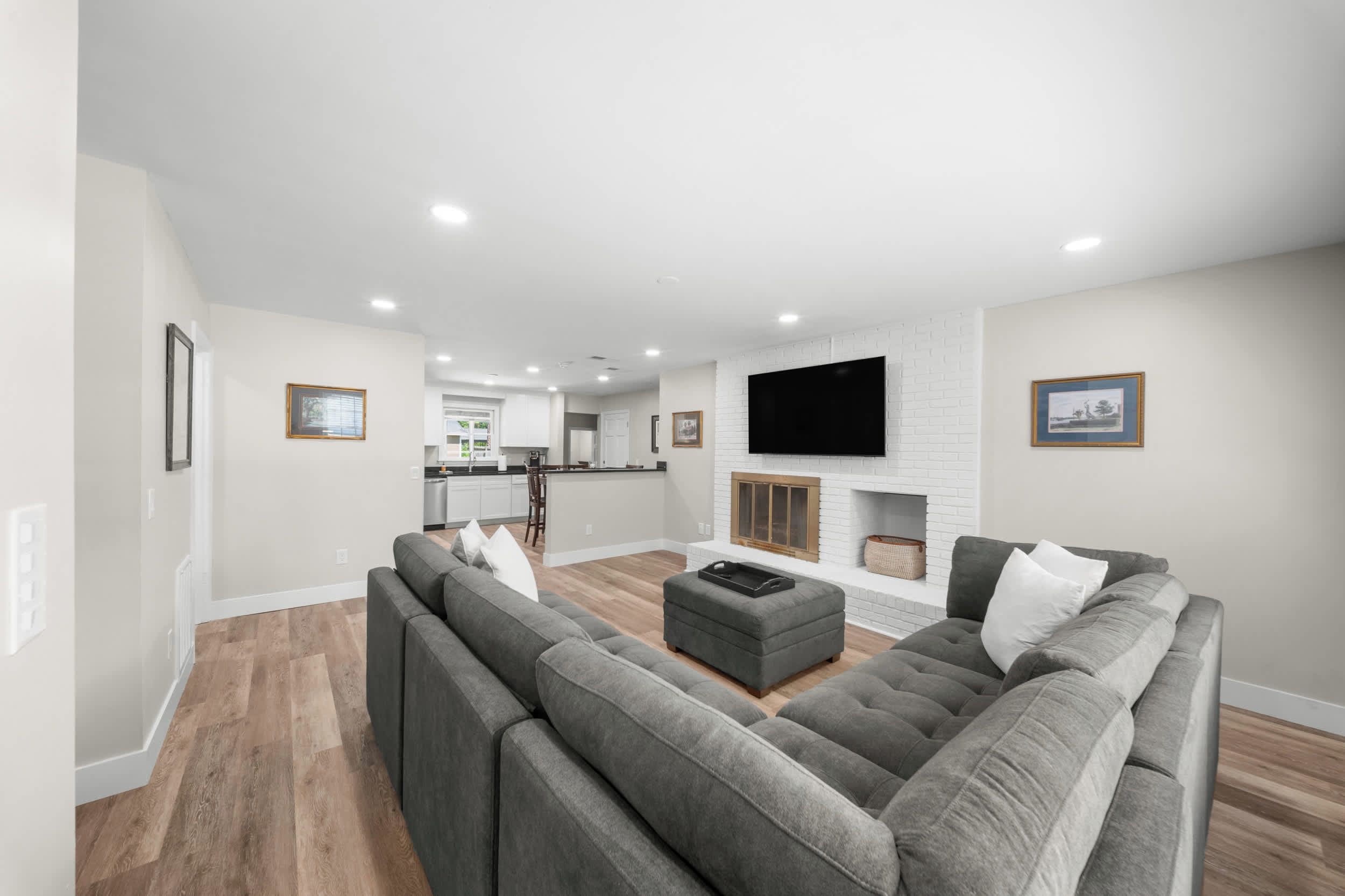 A modern living room with a grey sectional sofa, a flat-screen TV mounted on a white brick wall, and a kitchen in the background.