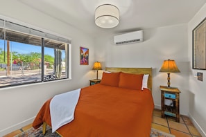 A bedroom with a large bed, two bedside lamps, an air conditioning unit, a painting, and a view of the outdoors through a window. - Casita Anita Historic Adobe Near Downtown Tucson (Tucson)