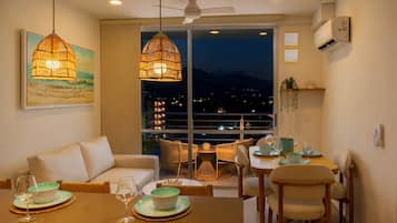 A dining area with a table set for two, a sofa, and a balcony with a view of the city at night.