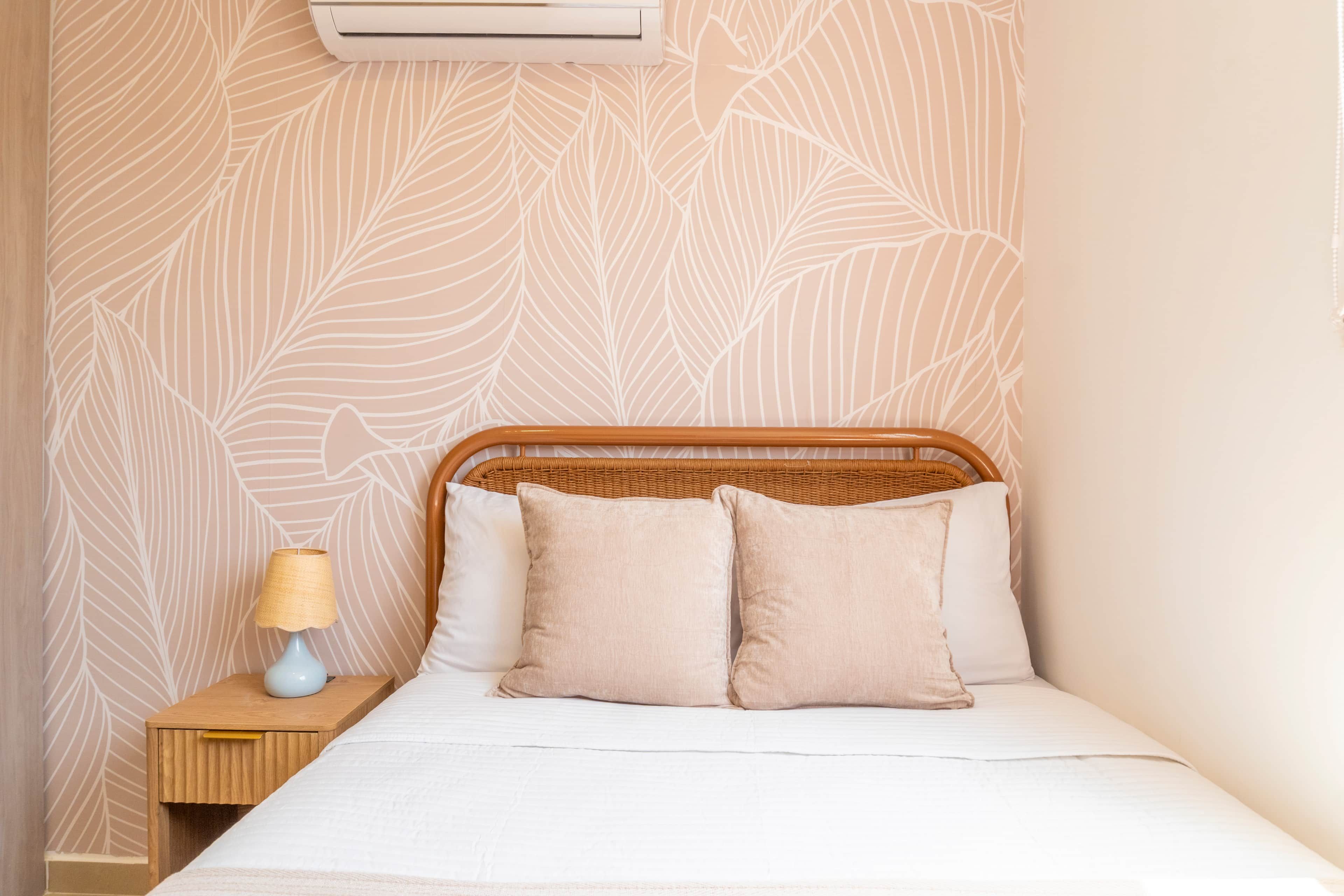 Un lit avec une tête de lit, deux oreillers, une table de chevet avec une lampe et un mur orné d’un motif de feuilles.