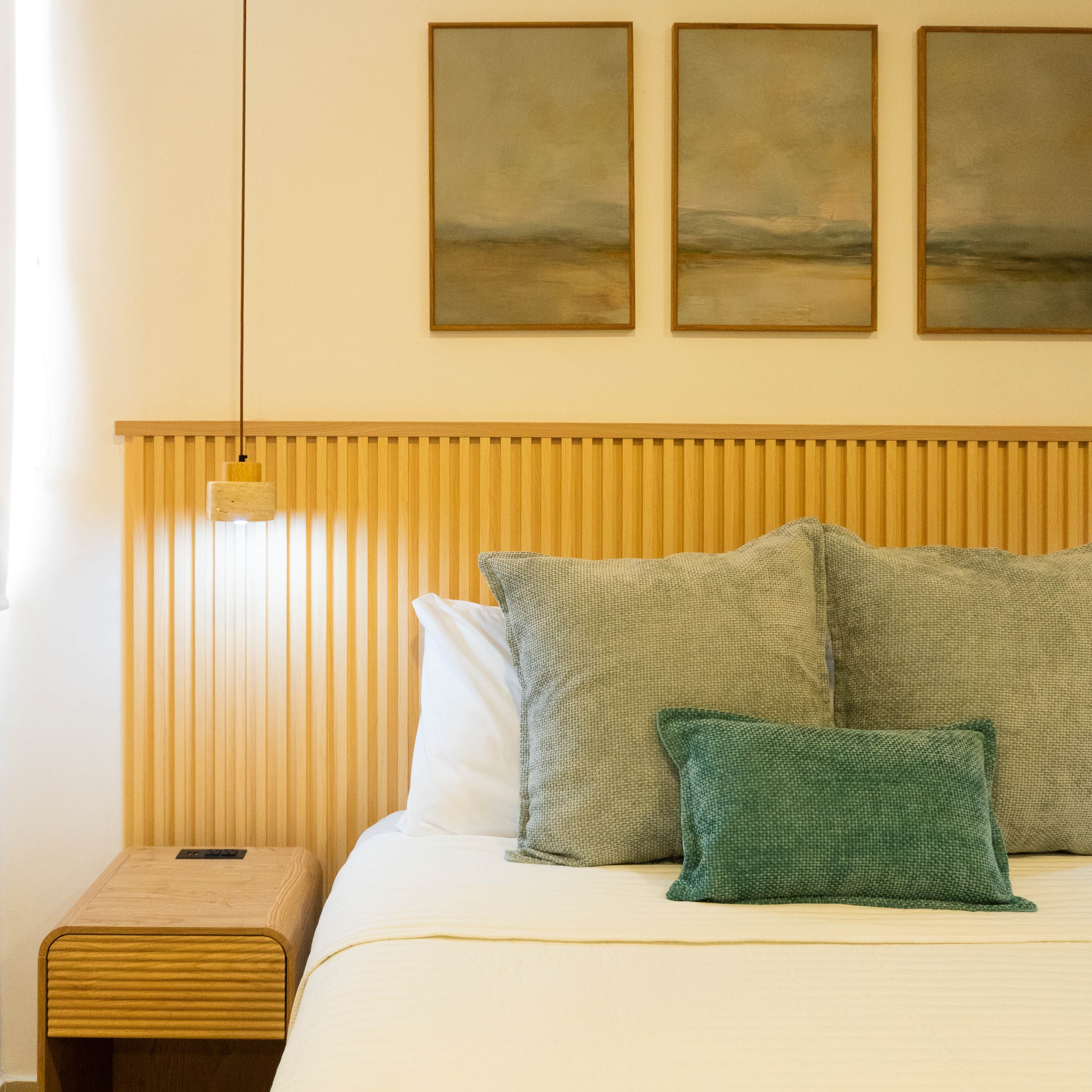 A neatly made bed with a wooden headboard, a bedside table, and three framed artworks on the wall.