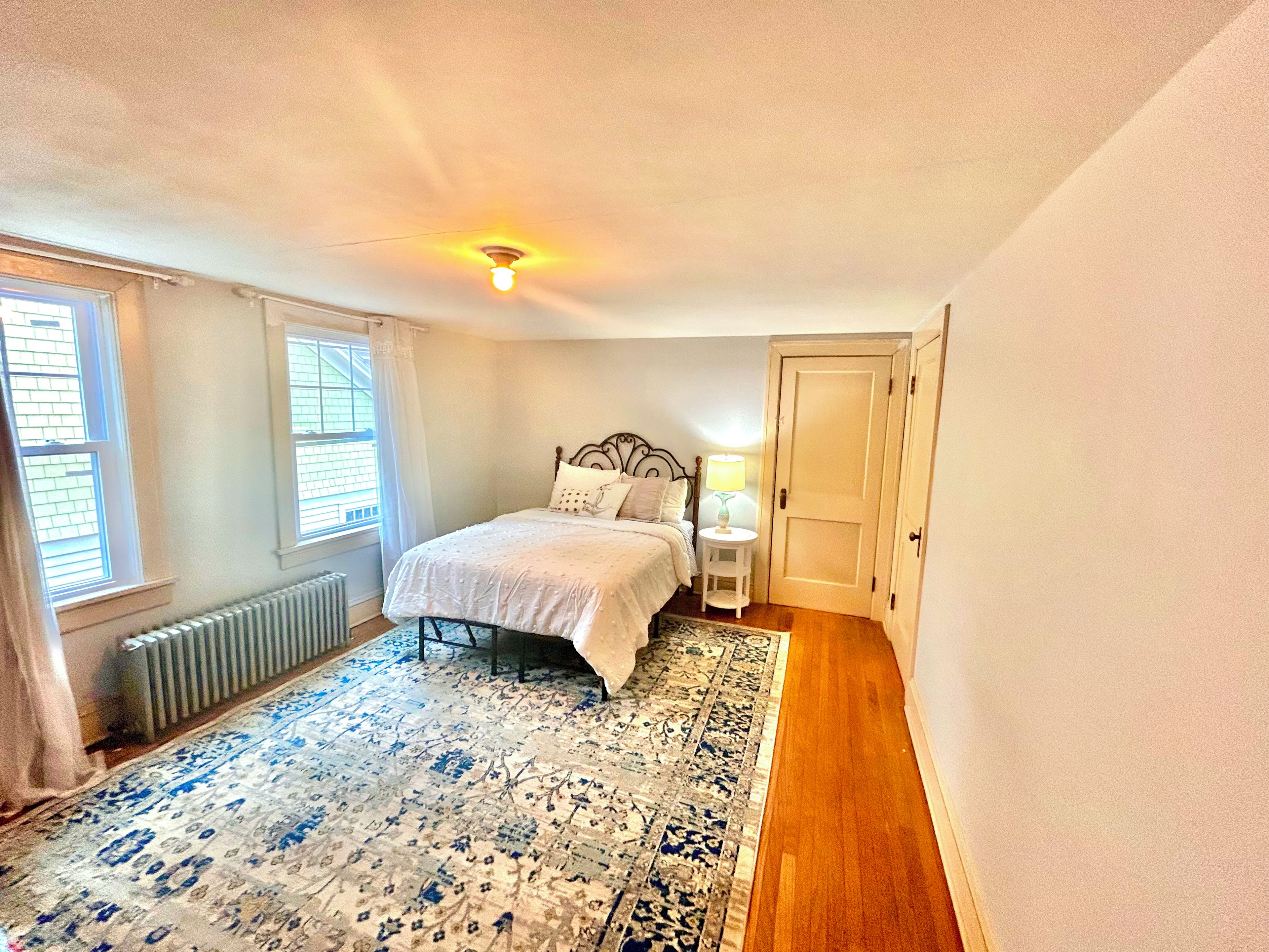 Une chambre à coucher avec un lit, un radiateur, une fenêtre avec des stores, une porte et un tapis.