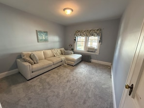 A living room with a beige sectional sofa, a radiator, a window with curtains, and a ceiling light fixture. - Classic Cape with Modern Cottage Charm (Horseheads)