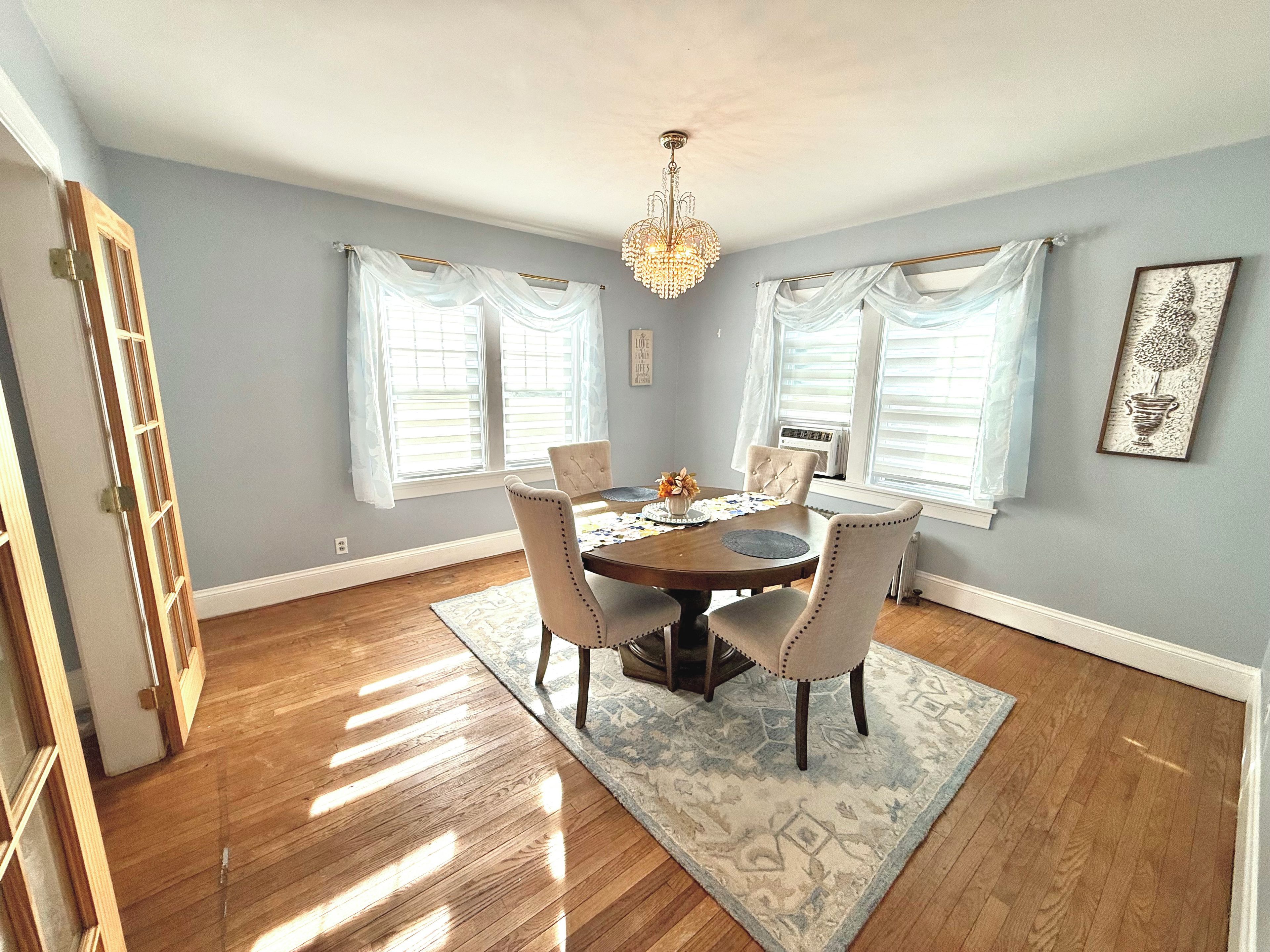 A dining room with a wooden table, four chairs, a chandelier, and large windows with white curtains.