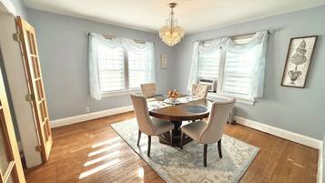 A dining room with a wooden table, four chairs, a chandelier, and large windows with white curtains.