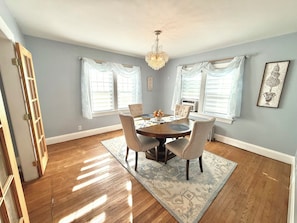 A dining room with a wooden table, four chairs, a chandelier, and large windows with white curtains. - Classic Cape with Modern Cottage Charm (Horseheads)
