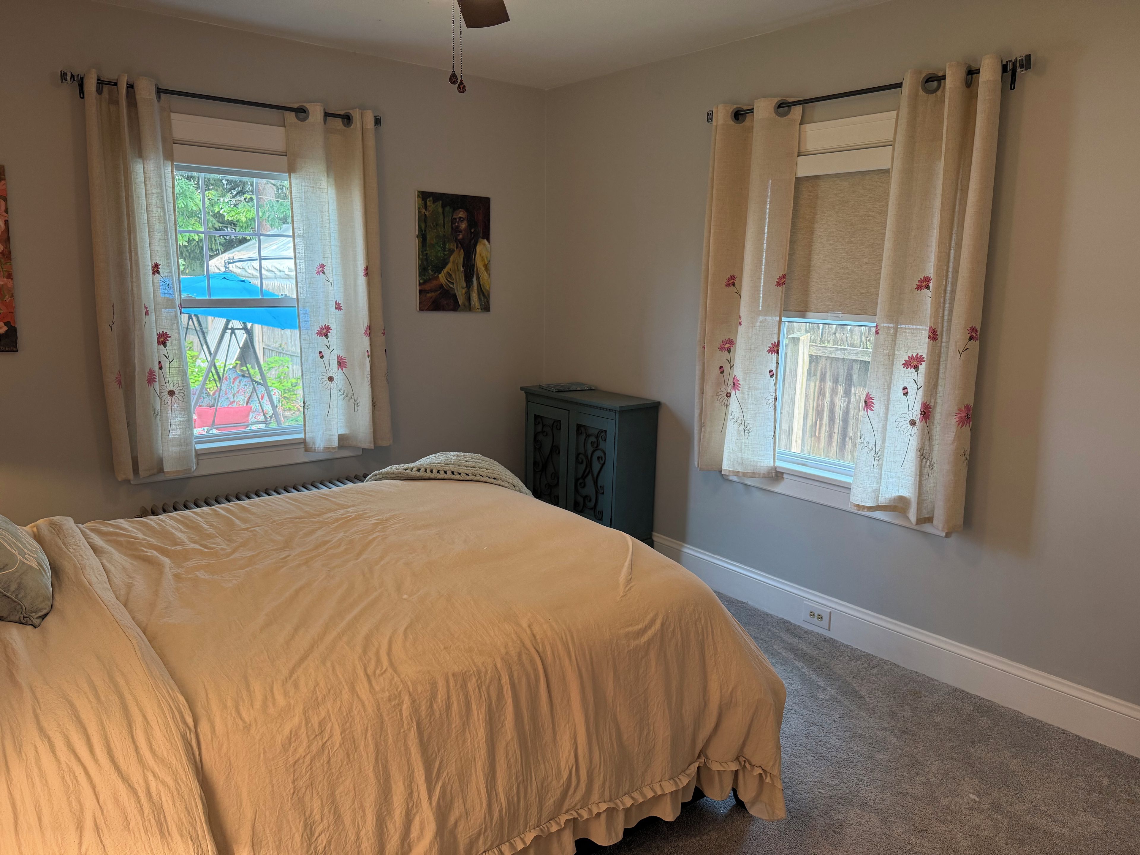 Quarto com cama, duas janelas com cortinas, radiador, pequeno armário e ventilador de teto.