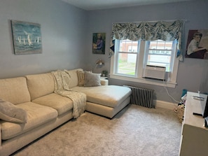 Un salon avec un canapé beige, un radiateur, une fenêtre avec des rideaux et un tableau accroché au mur.